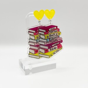 Peut inclure: Une paire de boucles d'oreilles en forme de cœur jaune avec une pile de livres colorés suspendus à chaque cœur. Les livres portent des mots comme "Rire", "Inspirer", "Sourire", "Grandir", "Enseigner", "Lire" et "Être gentil".