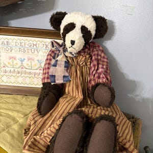 May include: A handmade panda bear doll with a white and black head, brown fur ears, and brown paws. The doll wears a red and brown striped dress and a blue and red patterned scarf. The doll is sitting on a yellow surface.