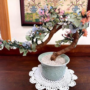 May include: A decorative bonsai tree sculpture featuring a textured brown trunk and branches adorned with translucent carved stone flowers in shades of pink, blue, and white, set in a pale green ceramic pot filled with small white pebbles.