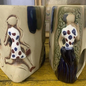 May include: Two ceramic mugs with a beige base color. Each mug features a raised relief of a figure in a dress with blue and red polka dots. The figures are set against a background of green and blue accents. The mugs have a unique, angular shape.
