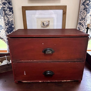 May include: A two-drawer wooden storage box with a dark red finish. The box has black metal handles and is sitting on a wooden surface. A framed picture and a window with curtains are in the background.