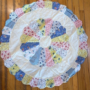 May include: A circular fabric mat featuring a central ring of multi-colored floral and checkered fabric petals, a wide white circular border, and an outer scalloped edge composed of various patterned fabric segments in blue, yellow, and pink.