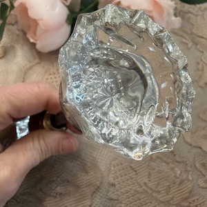 May include: A clear, faceted glass door knob with a brass base. The knob has a floral design and is held by a hand. The background includes pink flowers and a patterned fabric.