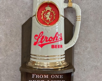 May include: Vintage Stroh's Beer stein-shaped wall decor. The white ceramic stein features a gold-colored lid and handle, with a red and gold emblem. The words "Stroh's BEER" are in red. A wooden plaque below reads "FROM ONE BEER LOVER TO ANOTHER."