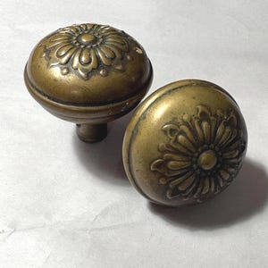 May include: Two antique brass door knobs. Each knob has a rounded shape with a floral design on the front. The knobs have a warm, aged patina, suggesting they are vintage or antique. The knobs are likely for a door or cabinet.