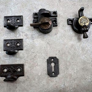 May include: A collection of dark metal hardware including three rectangular sash lifts with fleur-de-lis motifs, two window sash locks with circular floral accents, and one vertical keyhole escutcheon plate with two screw holes for mounting.