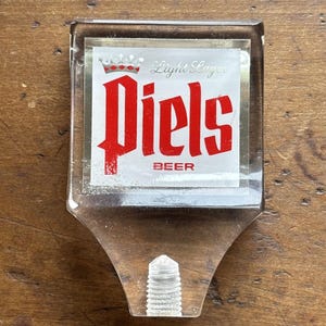 May include: Clear plastic beer tap handle with the word "Piels" in red block letters. The handle has a rectangular top with the words "Light Lager" and a crown design. The bottom of the handle has a screw-like design.