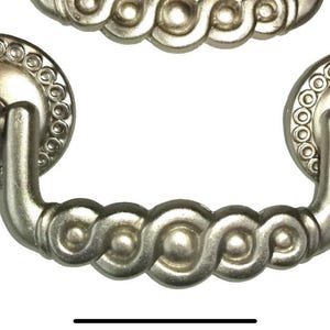 May include: Silver-toned cabinet pull with a decorative design. The pull features a circular backplate with a beaded edge and a curved handle with a repeating swirl pattern. Hardware for furniture.