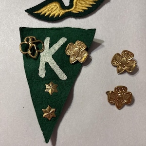 May include: A collection of Girl Scout memorabilia, including a green felt pennant with a white "K" and gold stars, along with several gold-colored pins and a green and gold embroidered wing patch. The items are arranged on a white surface.