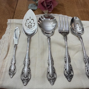 May include: A set of five silver-toned serving utensils featuring ornate floral embossed handles. The collection includes a small spreader, a large pierced cake server, a wide scalloped bowl spoon, a three-tined fork, and a pierced serving spoon.