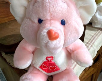 May include: A plush toy featuring a light pink bear with large white ears, blue eyes, and a heart-shaped orange nose. The bear has a white belly patch displaying a red graphic of a weight with two small white hearts inside the base of the weight.