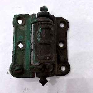 May include: An antique metal hinge with a dark brown finish and green patina. The hinge has a decorative finial and visible screw heads. The metal shows signs of age and wear, with a weathered appearance. The hinge has multiple screw holes.