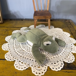 May include: A plush frog toy with olive green fur, a white underbelly, and white patches above its dark eyes. The stuffed animal is positioned in a prone, outstretched posture with its four limbs extended outward on a white crocheted doily.