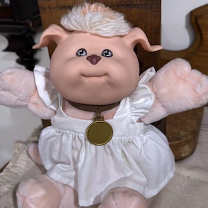 May include: A plush doll with light peach skin, floppy ears, and a tuft of light-colored yarn hair. The doll features a brown collar with a round gold-colored pendant and wears a white sleeveless dress with ruffled shoulder straps and a gathered skirt.