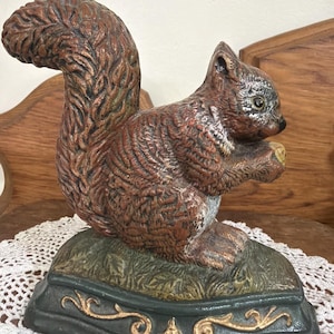 May include: A cast metal squirrel figurine featuring a textured brown coat and a large, bushy tail. The squirrel is sitting on a dark green base with gold-colored scrollwork details, holding a small nut in its front paws with a focused expression.