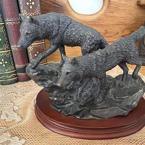 May include: A detailed sculpture of two wolves in a dynamic pose, crafted in a dark, metallic finish. The wolves are depicted on a rocky base, mounted on a round, dark wooden stand. The sculpture is a decorative item.