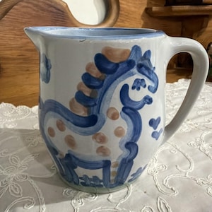 May include: White ceramic pitcher with a blue rim and handle. The pitcher features a hand-painted design of a horse with a blue mane and tail, and brown spots. The design includes blue hearts and floral accents.