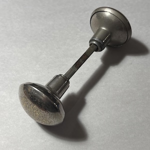 May include: A vintage silver-toned door knob with a textured, rounded handle. The knob has a long, threaded stem connecting to a smaller, circular base. The metal shows signs of age and wear, with a slightly tarnished appearance.