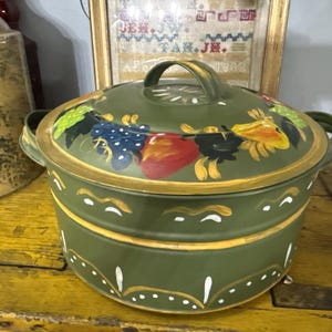 May include: A green ceramic casserole dish with a lid, featuring a gold rim and handle. The lid is decorated with colorful painted fruits and leaves. The dish has a two-tiered design and is set on a yellow wooden surface.