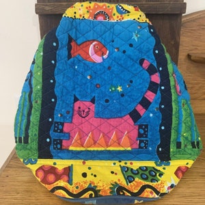 May include: Quilted tea cozy with a whimsical design. Features a pink cat, orange fish, and a blue background with stars. The cozy has a yellow and blue patterned border and a quilted texture.