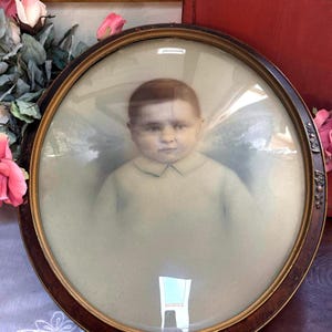 May include: An oval-shaped portrait frame with a dark wood finish and a gold inner rim, featuring a convex glass cover. Inside the frame is a sepia-toned photograph of a young child with short hair wearing a collared shirt, set against a soft background.