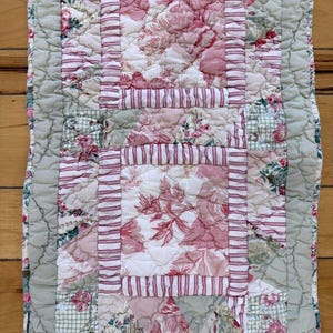 May include: A quilted table runner with a patchwork design. The runner features a central panel with floral patterns in shades of pink and white, framed by striped and solid fabric sections. The edges are a soft green, creating a vintage aesthetic.