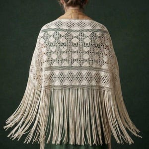 May include: An off-white crochet shawl featuring a geometric diamond pattern and a wide border of long, straight fringe. The garment is draped over the shoulders, showcasing intricate openwork stitching and a tan collar detail at the neckline.