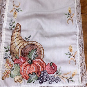 May include: A white rectangular table runner featuring a cross-stitched cornucopia overflowing with an orange pumpkin, red apple, green pear, and purple grapes. The fabric is bordered by white lace trim and accented with small wheat stalks.
