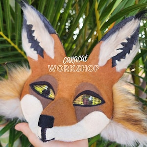 Può includere: Un primo piano di una maschera di volpe marrone e bianca con orecchie nere e occhi in rete gialla. La maschera è realizzata in feltro e pelliccia. Il testo "caracal WORKSHOP" è stampato sulla maschera.