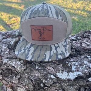 以下が含まれることがあります： 「Three Rivers Hat Co.」と書かれた茶色の革のパッチが付いた迷彩柄の野球帽
