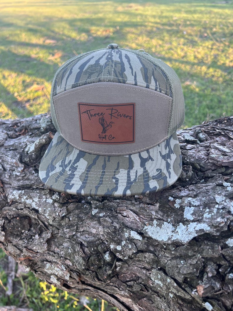 以下が含まれることがあります： 「Three Rivers Hat Co.」と書かれた茶色の革のパッチが付いた迷彩柄の野球帽