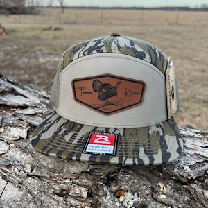 May include: A camouflage-patterned flat bill snapback hat with a brown leather patch featuring a turkey graphic and the words "Three Rivers". The hat has a mesh back and a "Flat Bill Snapback" tag.