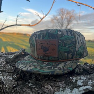 May include: A green and brown camouflage baseball cap with a brown leather patch that says "Three Rivers Hat Co."