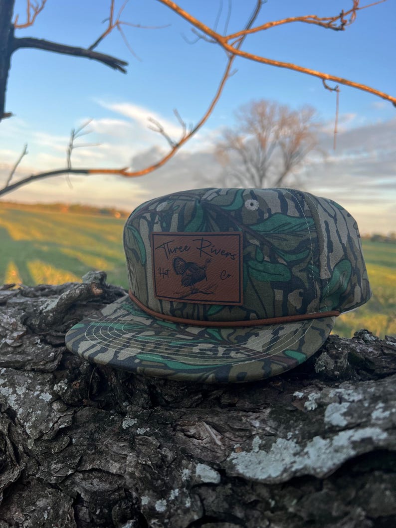 May include: A green and brown camouflage baseball cap with a brown leather patch that says "Three Rivers Hat Co."