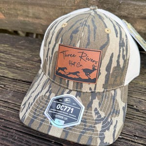 May include: A camouflage-patterned trucker hat with a white mesh back. The hat features a brown leather patch with the text "Three Rivers Hat Co." and a graphic of animals. A tag with the text "OC771" is attached.