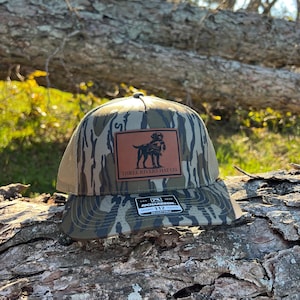 May include: A camouflage-patterned baseball cap with a brown leather patch featuring a dog graphic and the text "THREE RIVERS HAT CO." The cap has a mesh back and a curved brim. The Richardson brand tag is visible.