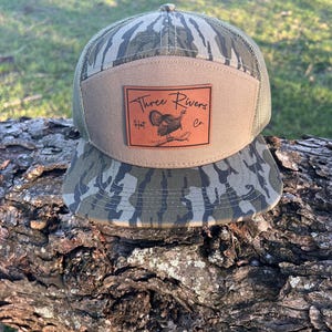 May include: A camouflage trucker hat with a brown leather patch that says "Three Rivers Hat Co." and features a turkey illustration.