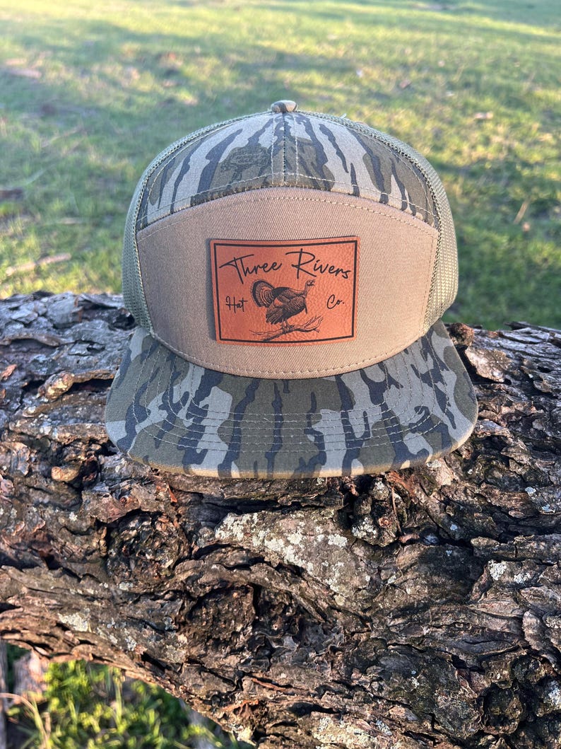 May include: A camouflage trucker hat with a brown leather patch that says "Three Rivers Hat Co." and features a turkey illustration.