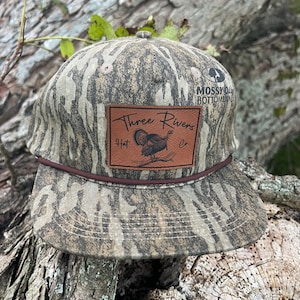 May include: A camouflage baseball cap with a brown leather patch that says "Three Rivers Hat Co." and features a turkey illustration. The cap has a brown brim and a Mossy Oak Bottomland logo on the side.