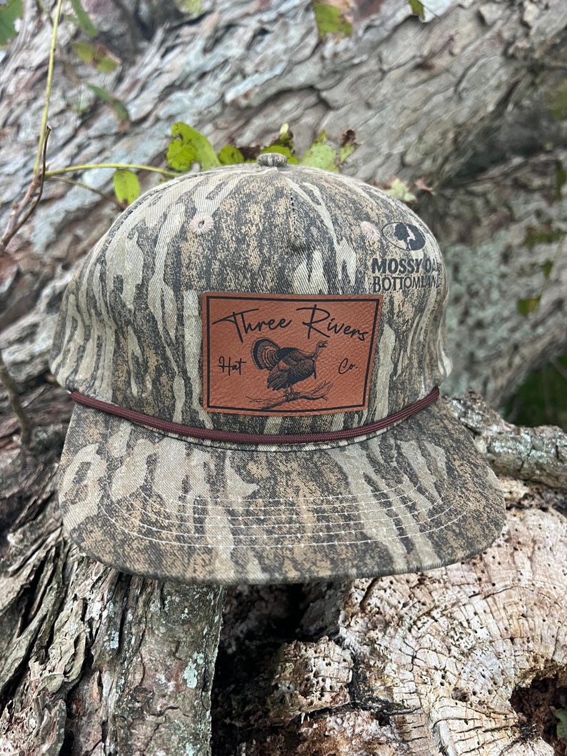 May include: A camouflage baseball cap with a brown leather patch that says "Three Rivers Hat Co." and features a turkey illustration. The cap has a brown brim and a Mossy Oak Bottomland logo on the side.