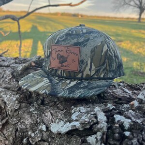 May include: A camouflage baseball cap with a brown leather patch that says "Three Rivers Hat Co." and features a turkey illustration.