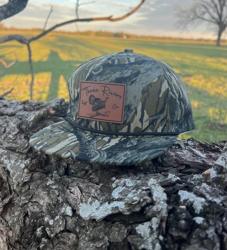 May include: A camouflage baseball cap with a brown leather patch that says "Three Rivers Hat Co." and features a turkey illustration.