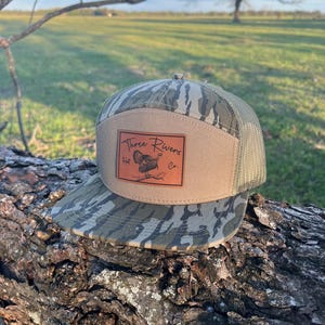 May include: A camouflage and brown mesh trucker hat with a brown leather patch that says "Three Rivers Hat Co.". The patch features a turkey illustration.