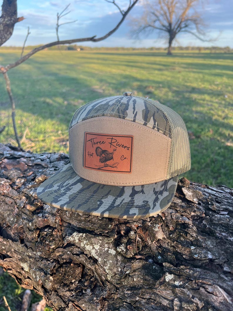May include: A camouflage and brown mesh trucker hat with a brown leather patch that says "Three Rivers Hat Co.". The patch features a turkey illustration.