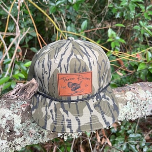 May include: A camouflage baseball cap with a brown leather patch that says "Three Rivers Hat Co.". The patch features a stylized image of a turkey.