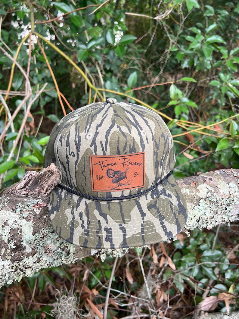 May include: A camouflage baseball cap with a brown leather patch that says "Three Rivers Hat Co.". The patch features a stylized image of a turkey.
