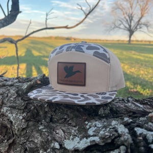May include: A tan and brown camouflage patterned baseball cap with a brown leather patch that says "Three Rivers Hat Co." and a silhouette of a duck flying.