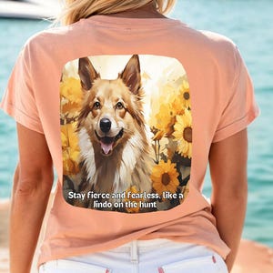 May include: Peach-colored t-shirt featuring a square graphic of a dog with sunflowers and the text: "Stay fierce and fearless, like a Jindo on the hunt." The dog has brown and white fur and is smiling.