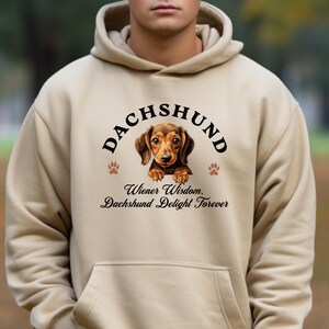 May include: A beige hoodie featuring a dachshund graphic. The graphic includes the word "DACHSHUND" above a brown and tan dachshund illustration, with the text "Wiener Wisdom, Dachshund Delight Forever" below.