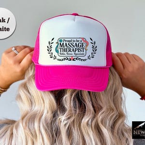 May include: A pink and white trucker hat with the text "Proud to be a MASSAGE THERAPIST" in a decorative frame. The hat has a pink brim and mesh back, and is worn by a person with blonde hair.